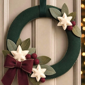 Può includere: Una ghirlanda circolare in feltro verde scuro con decorazioni che includono stelle in feltro color crema, piccole palline in feltro rosso scuro, foglie in feltro verde oliva con dettagli cuciti e un grande fiocco in feltro bordeaux.