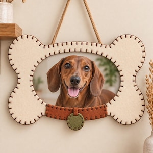 Puede incluir: Un marco de fotos de fieltro con forma de hueso en color crema con costuras en marrón oscuro. El marco presenta un detalle de collar de fieltro marrón en la parte inferior con una etiqueta circular verde oliva. Un perro salchicha marrón.