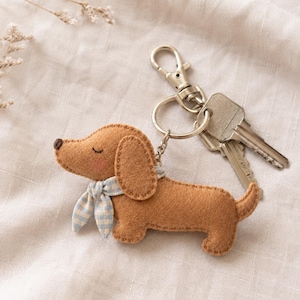 May include: A tan felt dachshund keychain featuring a light blue and white gingham neck scarf, a dark brown stitched nose, and closed eyes with pink cheeks. The dog figure is attached to a silver metal split ring and a lobster claw clasp.