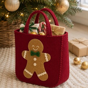 May include: A deep red felt tote bag featuring a tan gingerbread man appliqué on the front. The gingerbread man has a green bow tie, white stitched details, and two small buttons. The bag has two matching red felt handles and contains gifts.