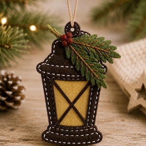 May include: A felt lantern ornament featuring a dark brown frame with white stitched detailing and a mustard yellow centre. The top is decorated with two green felt pine sprigs and three small red felt berries, attached to a tan hanging loop.