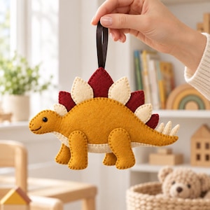 Puede incluir: Un adorno de fieltro con forma de estegosaurio en color amarillo mostaza con placas dorsales en crema y rojo oscuro. El dinosaurio presenta un pequeño ojo negro, costuras visibles en los bordes y una cinta marrón oscura para colgarlo.