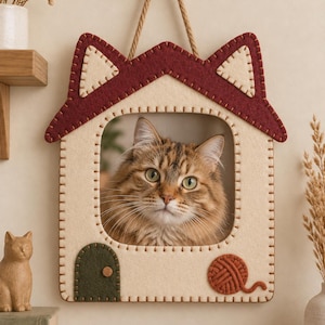 Può includere: Una decorazione da parete in feltro a forma di casa con una struttura color crema, un tetto rosso scuro, due orecchie color crema, una porta ad arco verde scuro e un gomitolo di lana arancione, che incornicia un gatto tigrato marrone.