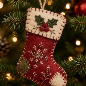 May include: A red felt Christmas stocking ornament featuring a cream-colored cuff with embroidered green holly leaves and red berries. The stocking body displays a white snowflake embroidery, a green heel patch, and a cream toe patch with stitching.