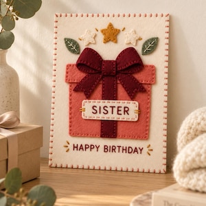 Puede incluir: Una tarjeta de cumpleaños rectangular de fieltro color crema con borde cosido. En el centro, un regalo rosa coral con un lazo rojo oscuro, dos hojas de fieltro verdes y tres estrellas. El texto SISTER y HAPPY BIRTHDAY está bordado.
