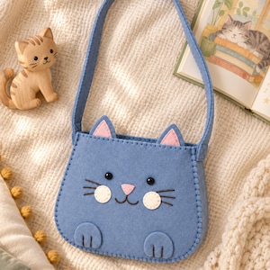 Puede incluir: Un bolso de mano de fieltro azul con forma de cara de gato, orejas rosas, ojos de cuentas negras, nariz rosa, mejillas blancas y dos pequeñas patas en la parte inferior. El bolso cuenta con una correa para el hombro y bigotes bordados en marrón.