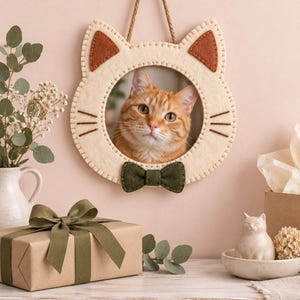 May include: A cream-colored felt cat-shaped picture frame featuring brown stitched ears, brown stitched whiskers, and a dark green felt bow tie at the bottom. The frame is suspended by a brown rope and contains a photograph of an orange tabby cat.