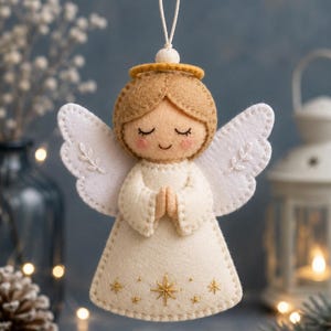 Peut inclure: Une décoration d'ange en feutrine avec une robe crème ornée d'étoiles brodées dorées, des ailes blanches avec des motifs de feuilles, et une tête beige avec une auréole dorée. L'ange a les yeux fermés, un sourire et des joues roses.