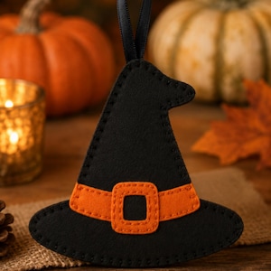 May include: A black felt witch hat ornament featuring an orange band and a square buckle detail. The hat is constructed with visible black stitching along the edges and includes a black ribbon loop at the top for hanging purposes.