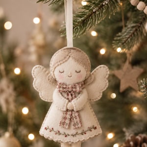 May include: A felt angel ornament in cream and beige tones, featuring stitched details, closed eyes, and rosy cheeks. The angel wears a floral patterned scarf and has a delicate embroidered vine with tiny red berries along the hem of the dress.
