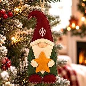 Pode incluir: Um ornamento de gnomo em feltro plano com um chapéu pontiagudo vermelho escuro com um floco de neve branco bordado, uma barba cor de creme, um corpo verde escuro e botas vermelhas. O gnomo segura uma estrela de feltro amarela brilhante.