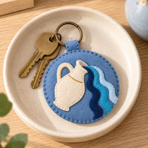 May include: A circular blue felt keyring featuring an embroidered cream-coloured pitcher pouring out three wavy streams of water in varying shades of blue. The keyring is attached to a bronze-toned metal ring holding two brass-coloured keys.