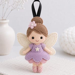 Può includere: Un ornamento a forma di fata in feltro con uno chignon castano decorato da un piccolo fiore rosa, ali color crema e un vestito color lavanda con una stella bianca. La figura ha un viso sorridente, scarpette da danza rosa e un nastro nero.