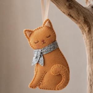 May include: A felt cat ornament in a warm orange hue, featuring stitched details, closed eyes, and pink cheeks. The cat wears a light blue scarf with a small white floral pattern and is suspended by a thin, light-colored ribbon loop for hanging.