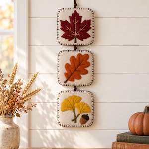 May include: A vertical wall hanging featuring three square cream-colored felt panels connected by dark brown cord. Each panel displays a different felt leaf appliqué: a deep red maple leaf, an orange oak leaf, and two yellow ginkgo leaves with an acorn.