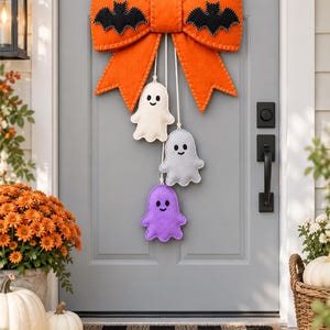 May include: A large orange felt bow decoration featuring two black felt bats attached to the top loops. Three small felt ghosts in cream, light grey, and purple hang from the centre of the bow by white cords, each with a stitched smiling face.