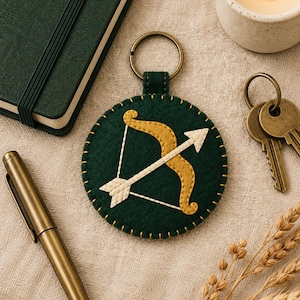 May include: A circular dark green felt keyring featuring an embroidered gold bow and a white arrow. The edges of the felt circle are stitched with gold thread, and it is attached to a circular bronze-toned metal ring at the top.