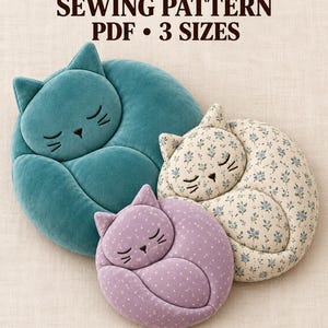 May include: A sewing pattern for a sleeping cat pillow in 3 sizes, 30, 40, and 50 cm. The image displays three finished pillows: one large teal velvet cat, one medium cream cat with a blue floral print, and one small lavender cat with white dots.