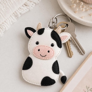 Puede incluir: Un llavero de fieltro con forma de vaca blanca y negra que presenta un hocico rosa, pequeños cuernos beige y ojos negros bordados. El llavero está unido a una anilla de metal plateado que sostiene dos llaves estándar, con costuras detalladas.