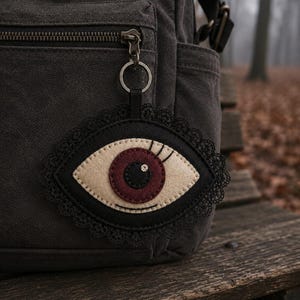 Op de afbeelding: Een donkergrijze canvas rugzak met een hangende bedel in de vorm van een oog. De bedel is gemaakt van vilt met een crèmekleurig oogwit, een donkerrode iris en een zwarte pupil, afgezet met een zwarte kanten rand en bevestigd aan een metalen ring.