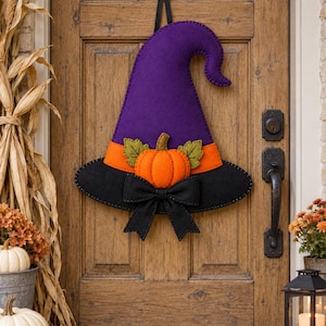 May include: A felt witch hat door hanging decoration featuring a tall purple cone, an orange band, and a wide black brim. The hat is embellished with a central orange pumpkin, two green felt leaves, and a black fabric bow at the base.