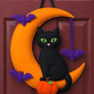 May include: A felt wall hanging featuring a large orange crescent moon, a black cat with bright green eyes, a small orange pumpkin with a green leaf, and three purple bats. The decoration is suspended by a black ribbon from a small black hook.