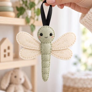 May include: A felt dragonfly ornament featuring a sage green body with stitched detailing, cream-colored wings, and small black bead eyes. The dragonfly has pink blush accents on its cheeks and a black ribbon loop attached to the top for hanging.