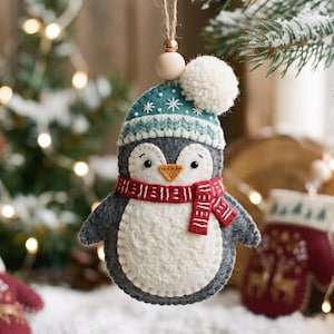 May include: A felt penguin ornament featuring a dark grey body, a white belly, and a teal winter hat with white snowflake embroidery and a white pom-pom. The penguin wears a red and white striped scarf and hangs from a jute string with a wooden bead.