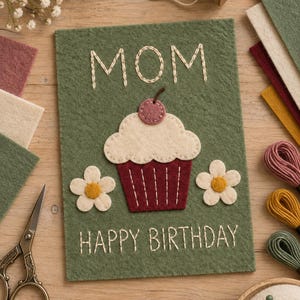 Può includere: Un biglietto rettangolare in feltro verde con il testo ricamato MOM e HAPPY BIRTHDAY in filo bianco. Al centro, un cupcake in feltro con base marrone scuro, glassa color crema e una ciliegia, affiancato da due fiori in feltro bianco.