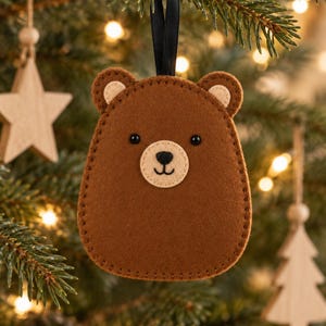 Puede incluir: Un adorno de fieltro marrón con forma de oso, con el hocico y el interior de las orejas de color crema, dos pequeños ojos de cuentas negras y una nariz y boca bordadas en negro. El adorno cuelga de una cinta negra fina.