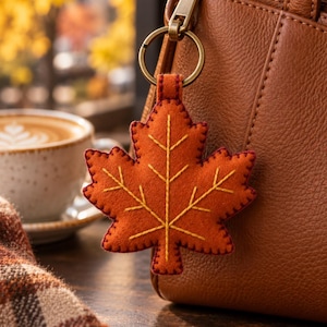 May include: A felt maple leaf keychain in a vibrant orange hue, featuring detailed yellow embroidered veins and dark red blanket stitching along the edges. The leaf is attached to a circular bronze-toned metal ring and clasp hardware.