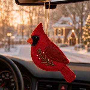 May include: A red felt cardinal bird ornament hanging from a string. The bird features a black face patch, an orange beak, and a stitched green pine branch detail with small white beads on its side. The felt is stitched along the edges.