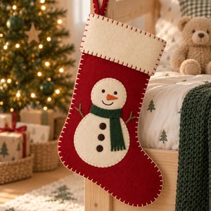 May include: A red felt Christmas stocking featuring a cream-coloured cuff and a central snowman appliqué. The snowman has a dark green scarf, brown stick arms, and three dark buttons. The stocking is finished with white blanket stitching.