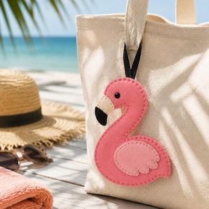 May include: A beige canvas tote bag featuring a pink felt flamingo ornament attached to the handle. The flamingo has a cream-coloured beak with a black tip, a small black eye, and a stitched wing detail, hanging from a thin black ribbon.