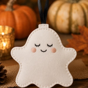 May include: A cream-coloured felt ghost ornament featuring embroidered closed eyes, a small smile, and soft pink cheeks. The ghost has a scalloped bottom edge and is topped with a thin black ribbon loop for hanging, with visible stitching along the perimeter.