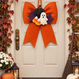 May include: A large orange felt bow hanging on a door, featuring a black felt pumpkin, two purple felt maple leaves, a white felt ghost, and two candy corn pieces attached to the center. The bow has long, pointed tails and a hanging loop.