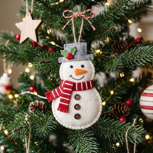 Könnte beinhalten: Ein Schneemann-Anhänger aus Filz mit einem grauen Zylinder, grünen Stechpalmenblättern und roten Beeren, einer orangefarbenen gestickten Nase, einem schwarzen Lächeln und einem rot-weiß gestreiften Schal. Drei braune Knöpfe sind mittig aufgenäht.