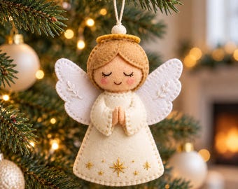 Pode incluir: Um enfeite de Natal de feltro em forma de anjo com vestido creme, cabelo castanho claro e asas brancas. O anjo tem olhos fechados, bochechas rosadas e mãos juntas em oração. O vestido tem estrelas douradas bordadas e uma auréola dourada.