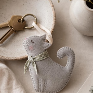 May include: A grey felt cat keyring featuring embroidered closed eyes, a pink nose, and stitched whiskers. The cat wears a small green floral patterned fabric scarf around its neck and is attached to a gold-toned metal ring by a cream ribbon.