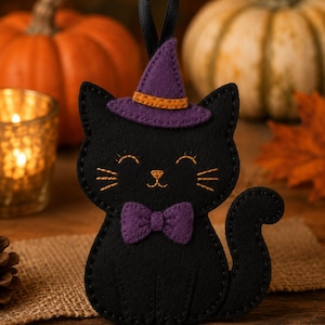 Può includere: Un ornamento in feltro nero a forma di gatto con un cappello da strega viola con fascia arancione e un papillon viola abbinato. Il gatto presenta occhi, baffi e naso ricamati in filo dorato, con un nastro nero in cima per appenderlo come decorazione.