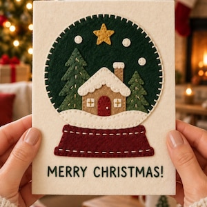 Può includere: Un biglietto d'auguri in feltro color crema con un'applicazione circolare a forma di palla di neve verde scuro con bordo cucito. All'interno si trova una casa color sabbia con porta rossa, tetto innevato, due pini verdi, una stella gialla.