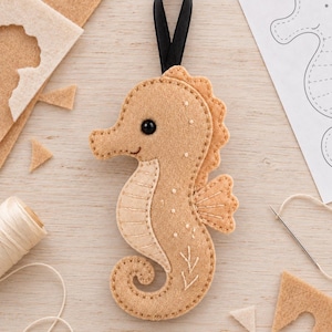 May include: A tan felt seahorse ornament featuring a black bead eye, stitched details on the belly and fins, and a black ribbon loop at the top for hanging. The seahorse has a curved tail and decorative white embroidered accents on its side.
