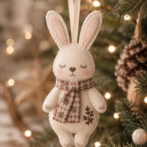 Könnte beinhalten: Ein cremefarbener Filz-Anhänger in Form eines Hasen mit langen Ohren, rosigen Wangen und geschlossenen Augen. Der Hase trägt einen gemusterten Schal mit roten und grünen Schneeflockenmotiven und hat einen gestickten Zweig mit roten Beeren.