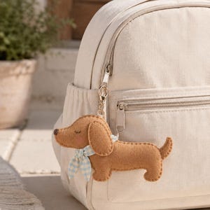 May include: A cream-colored canvas backpack featuring a brown felt dachshund keychain attached to the silver zipper. The dog-shaped charm has a light blue and white gingham patterned neckerchief, stitched details, and a small dark brown nose.
