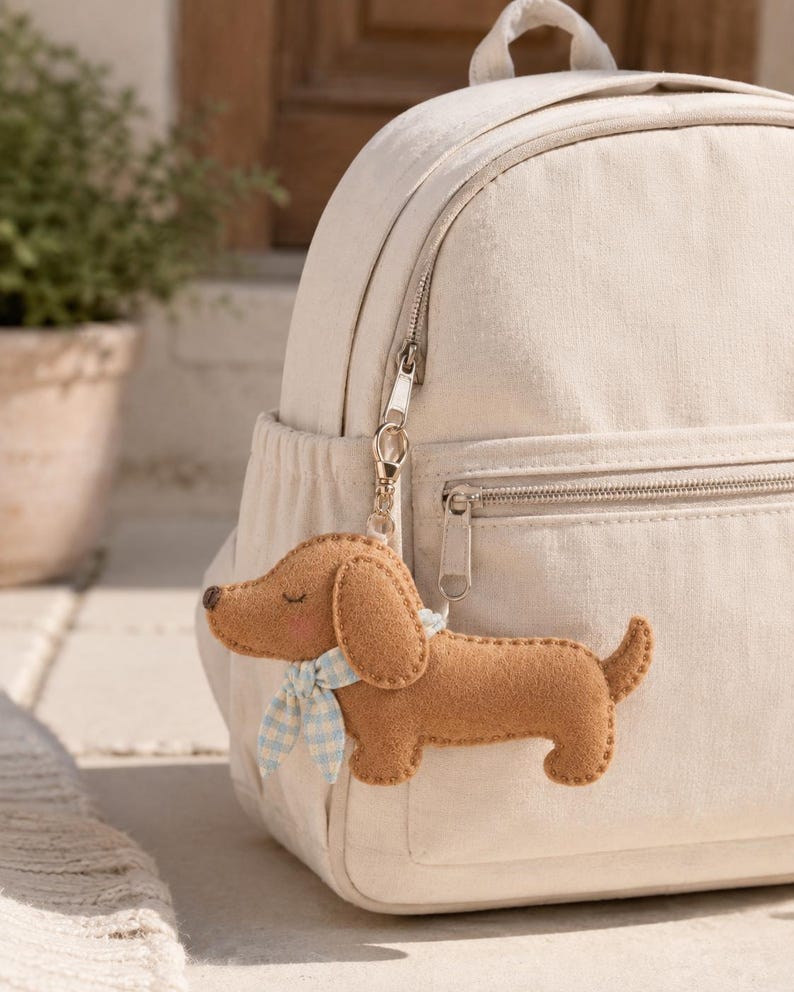 May include: A cream-colored canvas backpack featuring a brown felt dachshund keychain attached to the silver zipper. The dog-shaped charm has a light blue and white gingham patterned neckerchief, stitched details, and a small dark brown nose.