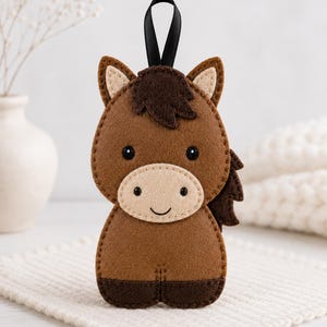 May include: A brown felt horse ornament featuring a beige muzzle, dark brown mane and tail, and black stitched eyes and mouth. The horse has a black ribbon loop at the top for hanging, with visible stitching along the edges of the felt pieces.