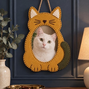 Puede incluir: Un marco de pared de fieltro color amarillo mostaza con forma de gato, cola verde oscuro, interior de orejas color crema y nariz marrón oscuro. El marco tiene una abertura ovalada central que muestra la foto de un gato blanco de ojos verdes.