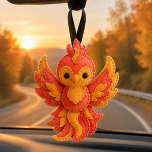 May include: A felt phoenix ornament in shades of orange and yellow with black bead eyes, featuring layered wing and tail feathers with visible stitching. The bird is suspended by a black ribbon loop, designed to hang from a rearview mirror.