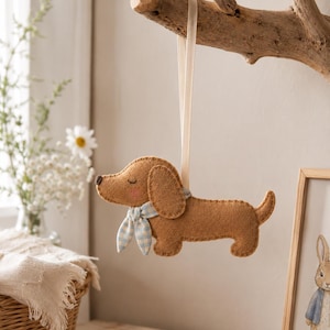 May include: A brown felt dachshund ornament featuring a light blue and white gingham patterned neckerchief, closed eyes, and pink cheeks. The dog is suspended by a thin cream-colored ribbon attached to its back, showcasing visible stitching.