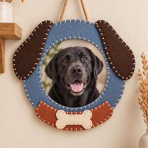 May include: A circular wall hanging frame featuring a blue felt border with white stitching, two dark brown felt dog ears at the top, and a light tan felt bone shape at the bottom. A photograph of a black Labrador dog is centered within the frame.